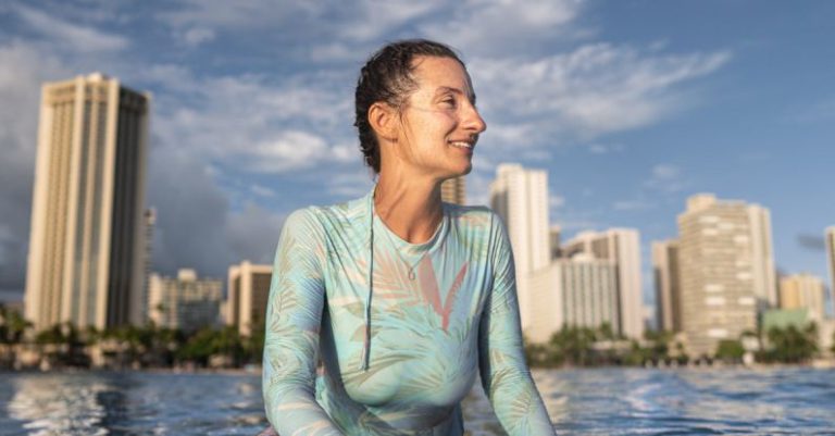 Surfing - Content young lady in wetsuit floating in sea sitting on surfboard while spending summer holidays in modern resort against blue sky