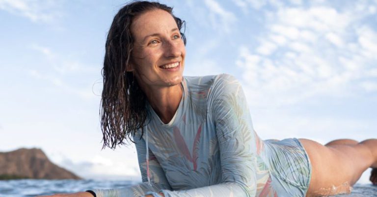 Surfing - Happy young female traveler with wet hair lying on surfboard and looking away after training in wavy ocean on sunny day